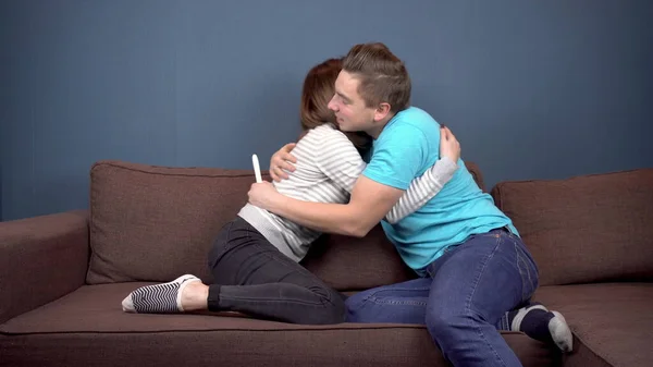 A young woman shows a positive pregnancy test to a young husband. A man rejoices and hugs his wife. Couple sit on the couch against each other — 스톡 사진
