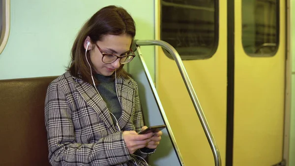 A young woman listens to music on headphones with a phone in her hands in a subway train. The girl is in correspondence on the phone. Old subway car