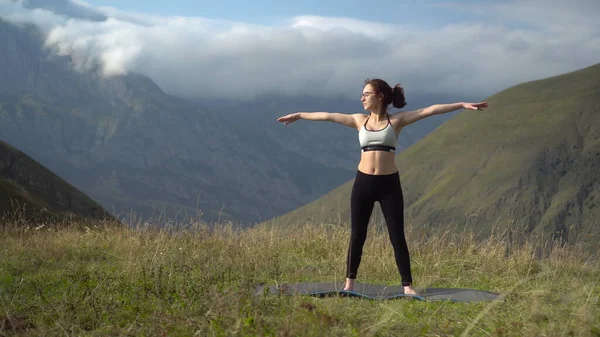 Mujer joven en chándal se dedica a realizar yoga triconasana pose en las montañas. La cámara se mueve para crear un efecto de paralaje. — Foto de Stock