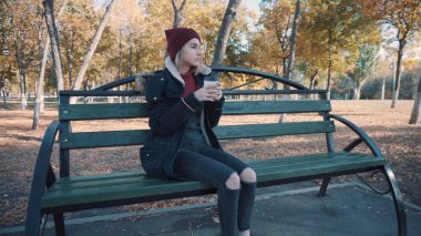 Kız parkta bir bankta kahve içiyor. Ceketli ve şapkalı kız.