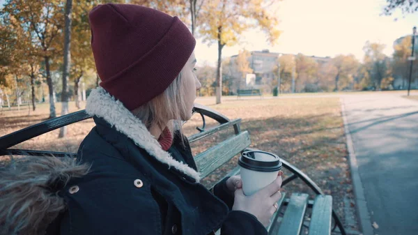 Chica bebe café en un banco en el Parque. Chica en una chaqueta y sombrero —  Fotos de Stock