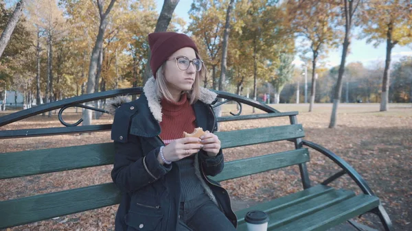 Una chica en un banco en el parque comiendo hamburguesa. Chica en una chaqueta y sombrero — Foto de Stock