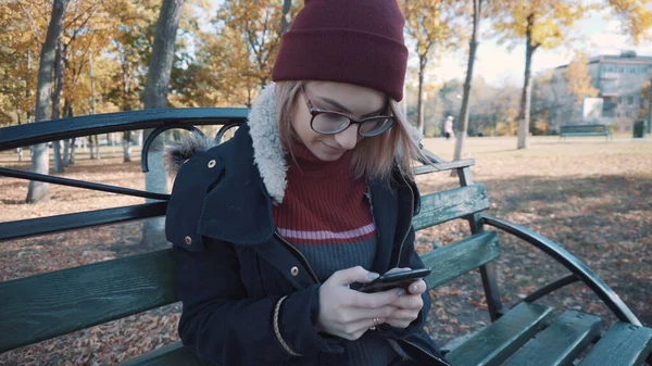 Chica sentada en un banco en el parque con tu teléfono. Chica en una chaqueta y sombrero — Foto de Stock