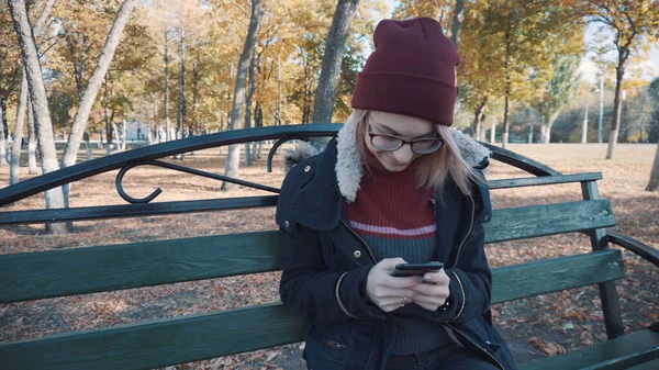 Egy lány ül a padon a parkban a telefonoddal. Lány zakóban és kalapban — Stock Fotó