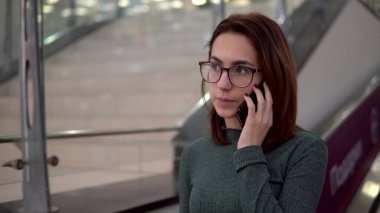 Genç bir kadın yürüyen merdivene biniyor ve telefonda konuşuyor. Büyük alışveriş merkezi
