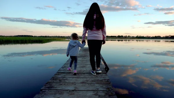 Mãe e filha caminham na ponte ao pôr-do-sol. Há um lago à volta da ponte. Nuvens refletidas no lago — Fotografia de Stock