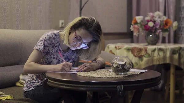 La chica escribe en las notas del cuaderno. Sentado en la sala de estar a la mesa —  Fotos de Stock