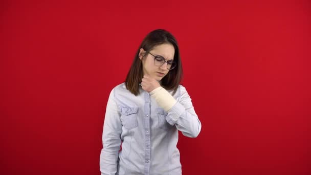 En ung kvinna med glasögon gör ont i armen och rör vid sin ömma arm. Fotografering på en röd bakgrund. — Stockvideo