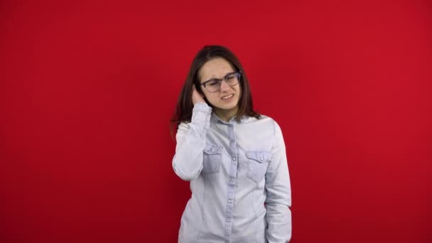 Une jeune femme avec des lunettes a mal à l'oreille et la tient dans ses mains. Tir sur un fond rouge. — Video