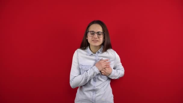 Een jonge vrouw met een bril heeft een hartzeer, ze houdt het vast met haar hand. Schieten op een rode achtergrond. — Stockvideo