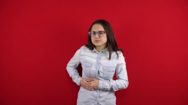 Gözlüklü genç bir kadının karaciğer ağrısı var, eliyle tarafını tutuyor. Kırmızı arkaplanda çekim.