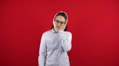 Gözlüklü genç bir kadının dişindeki ağrıdan dolayı başı geriye sarılmış. Kız eliyle yanağına dokunuyor. Kırmızı arkaplanda çekim.