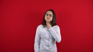 Gözlüklü genç bir kadının boğazı ağrıyor ve ona eliyle dokunuyor. Kırmızı arkaplanda çekim.