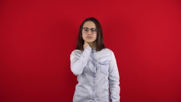 A young woman with glasses has a pain in her neck, she holds her neck with her hand. Shooting on a red background. — Stock Video