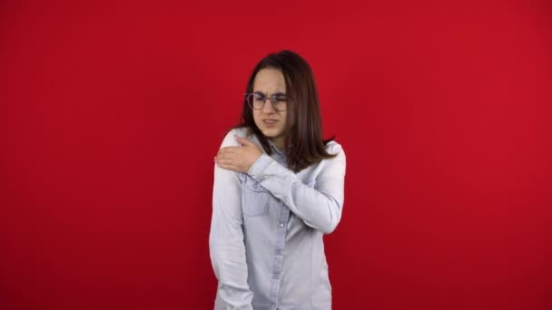 En ung kvinde i briller har en øm skulder, og hun rører den med hånden. Optagelse på en rød baggrund. – Stock-video