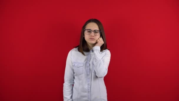 En ung kvinna med glasögon har tandvärk och rör vid kinden med handen. Fotografering på en röd bakgrund. — Stockvideo