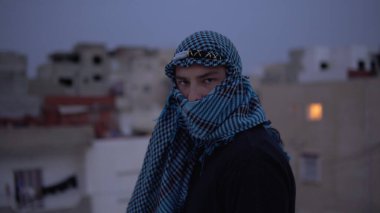 Arap mendili giymiş genç bir adam. Bir adam kameraya bakar ve yürür. Evin çatısında, Arap evlerinin arka planına karşı..