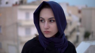 Arap şallı genç bir kadın. Bir kadın kameraya bakıyor. Evin çatısında, Arap evlerinin arka planına karşı..