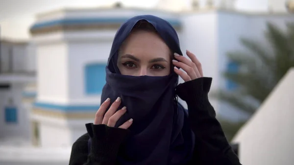 Young woman in arabic burqa. A woman looks at the camera and straightens a scarf. Against the background of the Arab house. — Stock Photo, Image