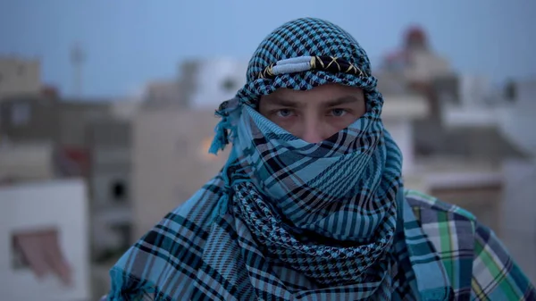 Un joven con un pañuelo árabe. El hombre mira a la cámara. En el techo de una casa sobre el fondo de las casas árabes. — Foto de Stock