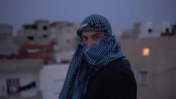 Молодой человек в арабском платке. Мужчина смотрит в камеру и идет. На крыше дома на фоне арабских домов. — стоковое фото