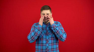 Genç adamın burnu ağrıyor, eliyle dokunuyor. Kırmızı arkaplanda çekim.