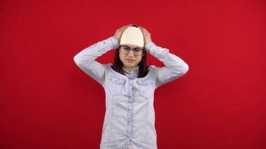 Genç kadının kafasında bir bandaj var, acıdan dolayı onu elleriyle tutuyor. Kırmızı arkaplanda çekim.
