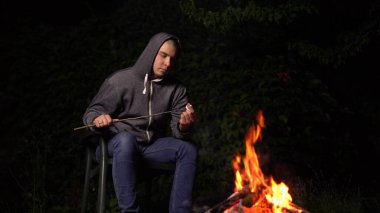 Kapüşonlu genç bir adam ateşin üstünde lokum kızartıyor. Geceleri kamp ateşi.