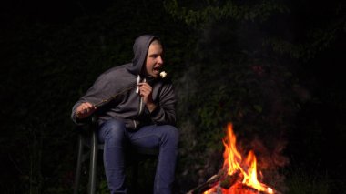 Kapüşonlu genç bir adam ateşin üstünde lokum kızartıyor. Geceleri kamp ateşi.