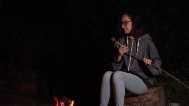 Kapüşonlu genç bir kadın ateşin üstünde marşmelov kızartıyor ve lokumlar alev alıyor. Geceleri kamp ateşi.