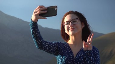 Elbiseli genç bir kadın dağlarda dikiliyor ve akıllı telefondan selfie çekiyor..