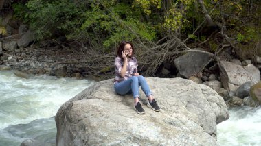 Genç bir kadın bir taşın üzerine oturur ve nehrin kenarındaki dağlarda telefonla konuşur..