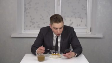 Takım elbiseli genç bir adam mısır gevreği yer, kahve içer ve akıllı telefon kullanır. Genç işadamı sabah mutfağında..