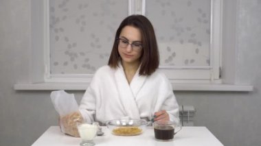 Beyaz önlüklü neşeli genç bir kadın mısır gevreği yer ve kahve içer. Sabah mutfağında gözlüklü genç bir kız..