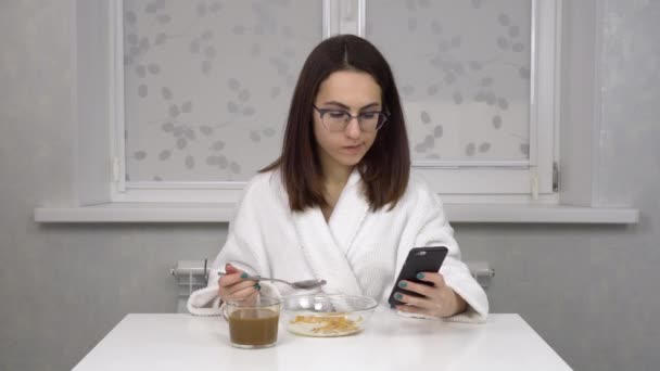 Wesoła młoda kobieta w białym płaszczu je płatki śniadaniowe, pije kawę i używa smartfona. Młoda dziewczyna w okularach w domu w kuchni rano. — Wideo stockowe