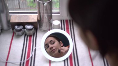 Pencerenin önündeki genç bir kadın küçük ışıklı bir aynaya bakar ve bir fırçayla makyaj yapar. Aynadan kıza bak..