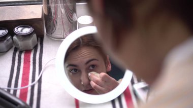 Pencerenin önündeki genç bir kadın ışıkla küçük bir aynaya bakar ve süngerle makyaj yapar. Aynadan kıza bak..