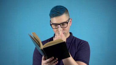 Mavi saçlı gözlüklü genç bir adam kitap okuyor. Alternatif bir adam. Elinde mavi bir arka plan olan bir kitap var..