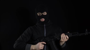 Kar maskeli bir adam AK-47 saldırı tüfeğiyle ayakta duruyor. Haydut makineye saldırır ve durur. Siyah bir arkaplanda.