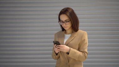 Genç bir kadın gri bir arkaplanda duruyor ve telefonda yazıyor..