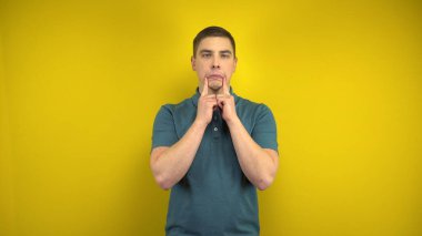 Genç bir adam, sarı arka planda parmaklarıyla onun yüzünde hüzünlü bir gülümseme yapıyor. Yeşil polo giyen adam..