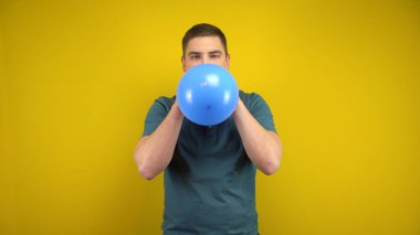 Genç bir adam sarı arka planda ağzı olan mavi bir balonu şişiriyor. Yeşil polo giyen adam..