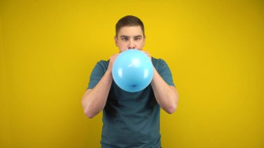 Genç bir adam sarı arka planda ağzı olan mavi bir balonu şişiriyor. Yeşil polo giyen adam..