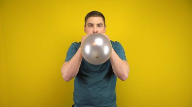 Genç bir adam sarı arka planda ağzı olan gümüş bir balonu şişiriyor. Yeşil polo giyen adam..