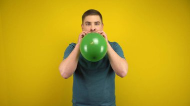 Genç bir adam sarı arka planda ağzı olan yeşil bir balonu şişiriyor. Yeşil polo giyen adam..