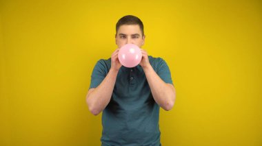 Genç bir adam sarı arka planda ağzı olan pembe bir balonu şişiriyor. Yeşil polo giyen adam..