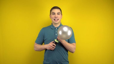 Genç bir adam sarı arka planda pompası olan gümüş bir balonu şişiriyor. Yeşil polo giyen adam..