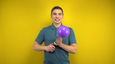 Genç bir adam sarı arka planda pompası olan mor bir balonu şişiriyor. Yeşil polo giyen adam..