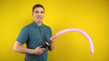 Genç bir adam sarı arka planda pompalı uzun pembe bir balonu şişirir. Yeşil polo giyen adam..