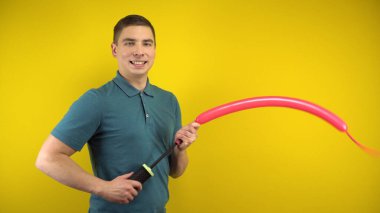 Genç bir adam sarı arka planda pompalı uzun kırmızı bir balonu şişirir. Yeşil polo giyen adam..